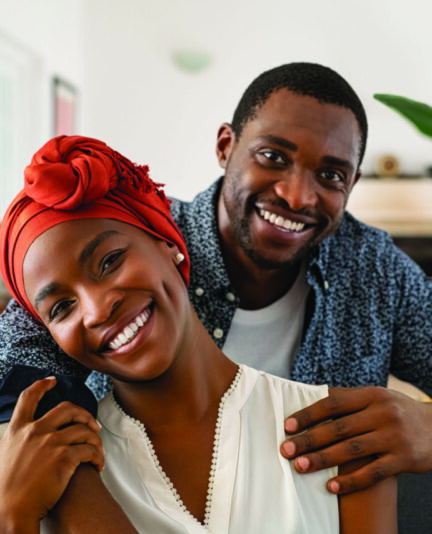 Portrait of mid adult happy african american couple hugging at home while sitting on couch. Portrait of mature man with his woman wearing traditional turban looking at camera. Black mid adult couple.